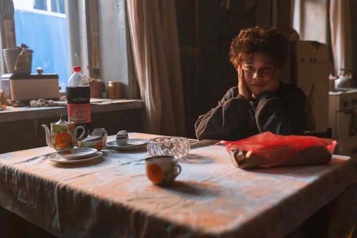 An image young Igor Grom (from Grom: Difficult Chilhood) sitting at a table in the morning, holding his head in his hands, and looking into the camera
