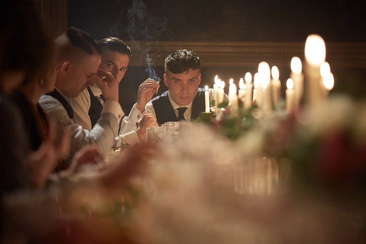 An image of Thomas Shelby (From Peaky Blinders) sitting at the end of a crowded dinner table smoking a cigarette
