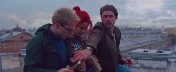 An screenshot of the movie Major Grom: Chumnoi Doktor, as three people sit on a rooftop. One of the characters (Igor Grom) reaches over the middle person to grab the other person's sketchbook