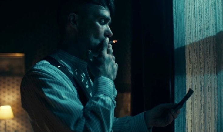 An screenshot of the show Peaky Blinders, as Thomas Shelby stands smoking in front of a blue window