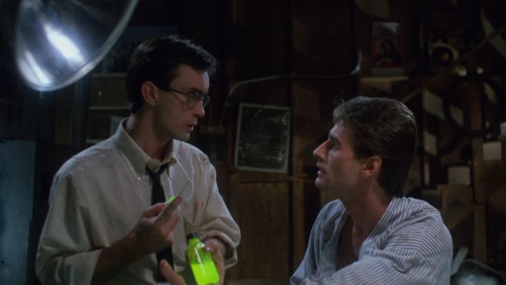 An screenshot of the movie Reanimator, as Herbert West and Daniel Cain sit in a dark basement while Herbert holds a glowing syringe