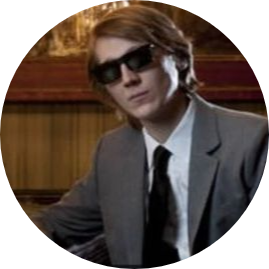 An image of Paul Dano sitting at a bar in a grey suit and glasses